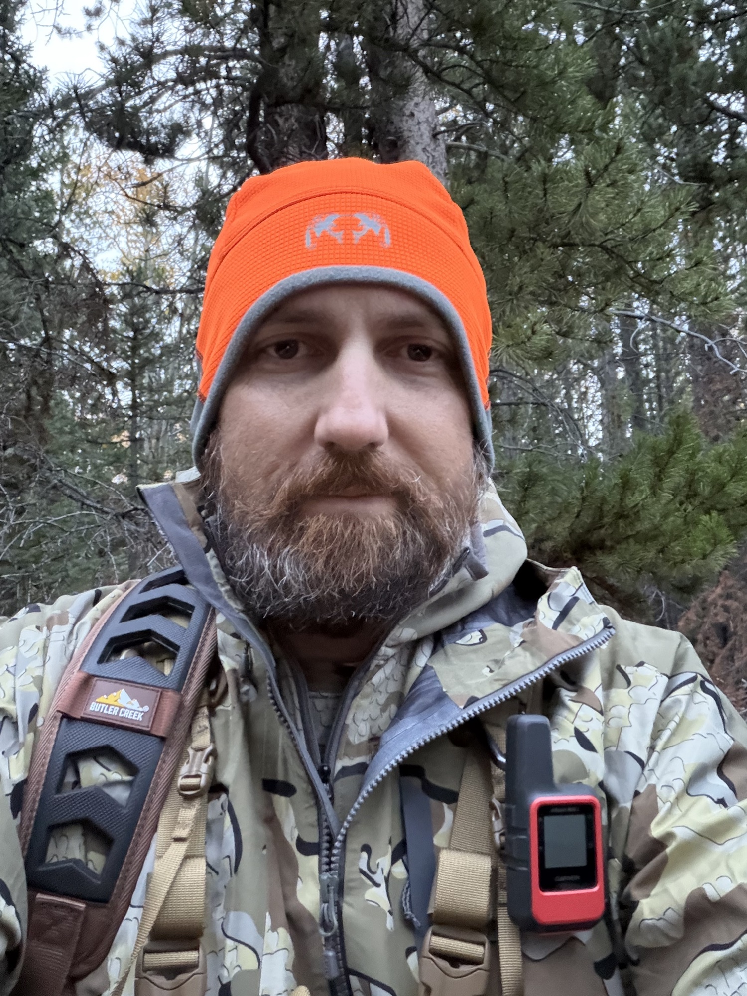John Mobley in the timber wearing a blaze orange beanie and camo jacket, with a Butler Creek sling and a Garmin inReach on his chest strap
