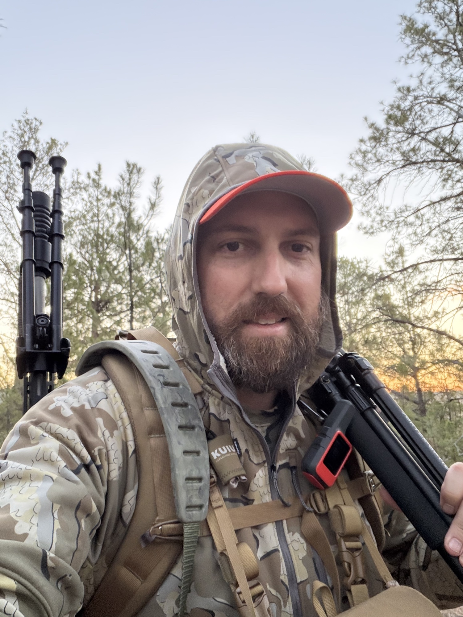 John Mobley in camo with a hood and orange-brimmed cap, carrying a pack with a Garmin inReach clipped to the strap, a bipod, and a tripod