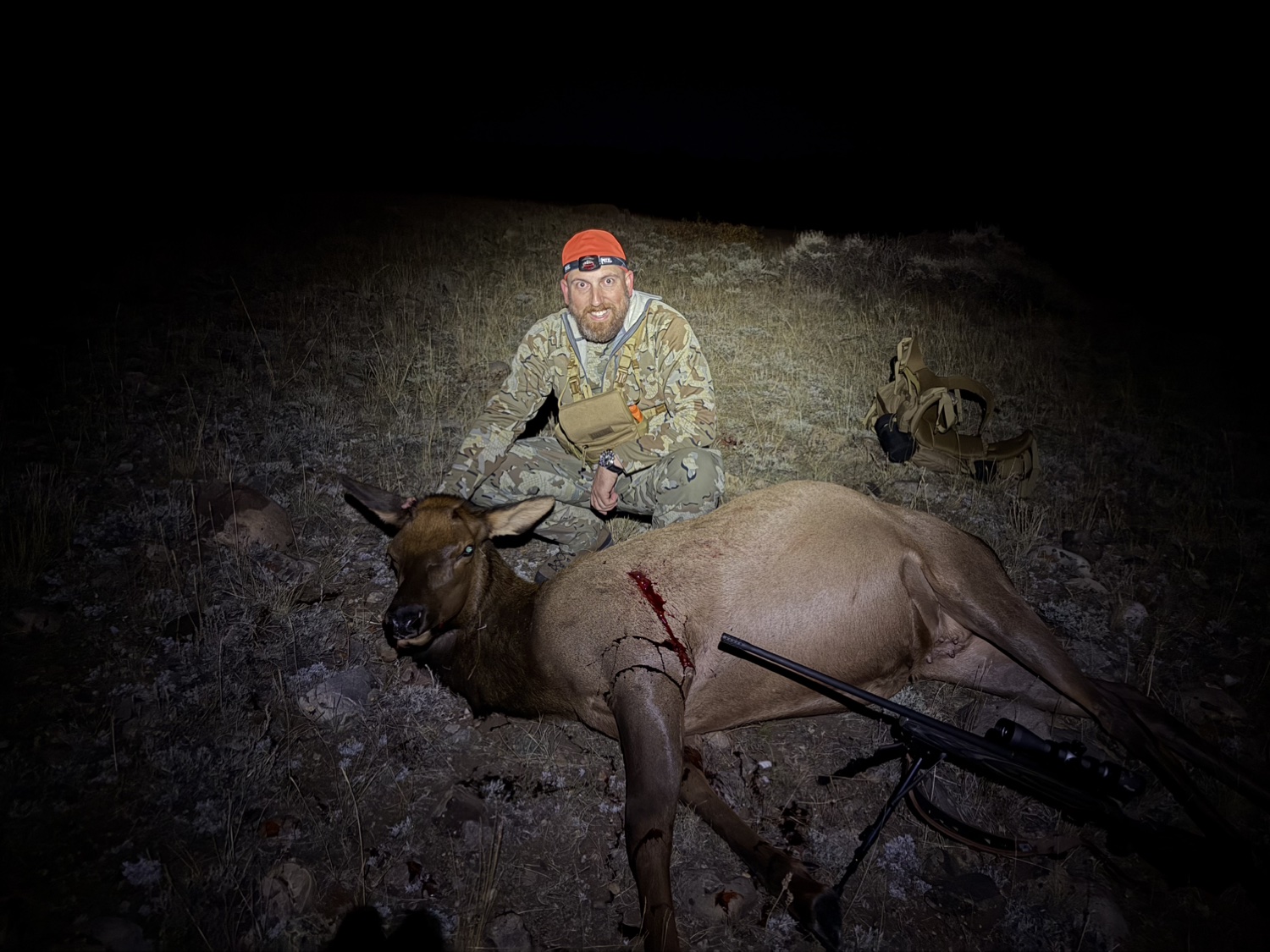 John Mobley at night with a harvested cow elk, his rifle resting on a bipod in front