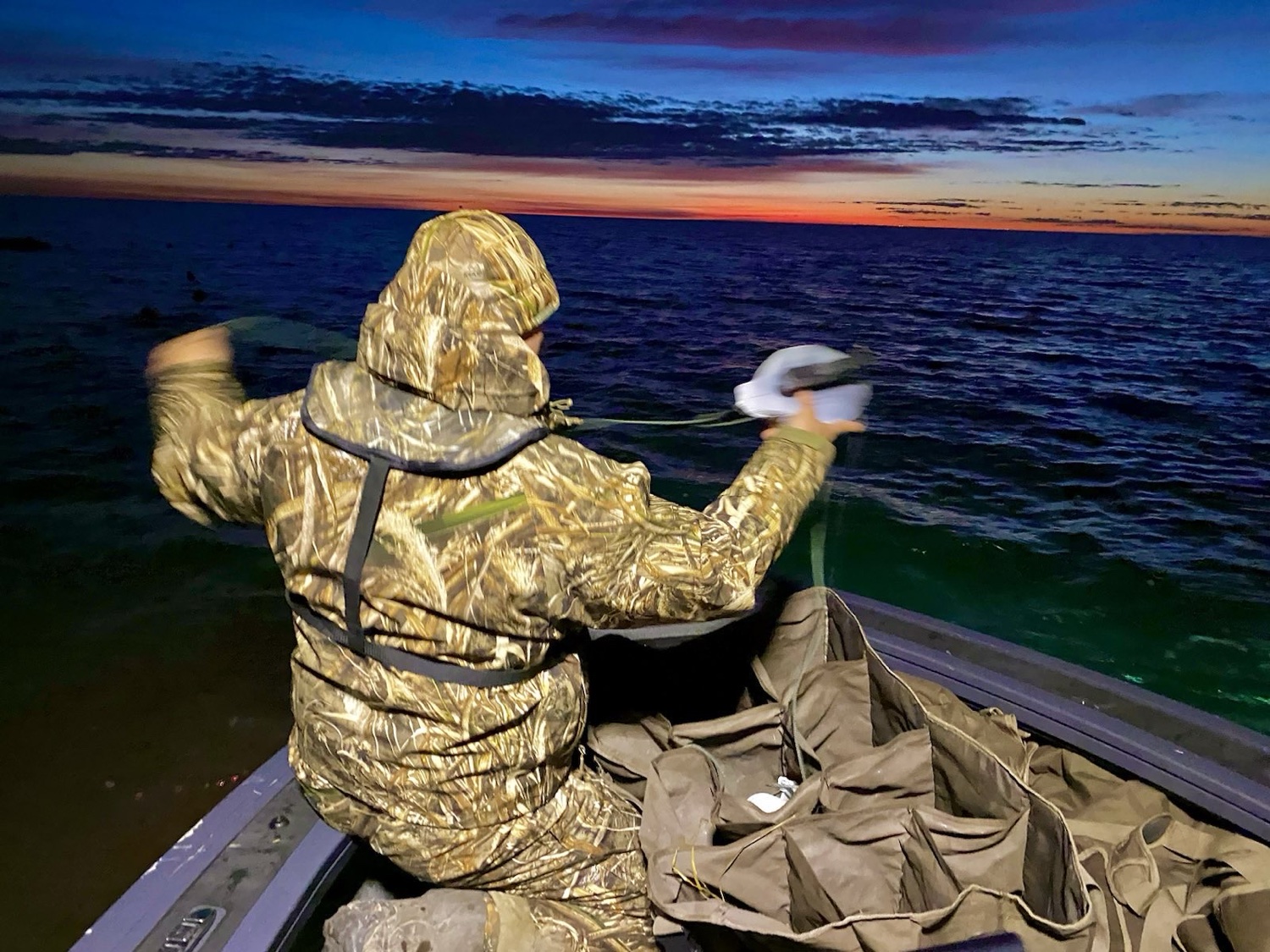 John Mobley in Realtree Max-5 camo setting out duck decoys from a boat at dawn, with a slotted decoy bag beside him