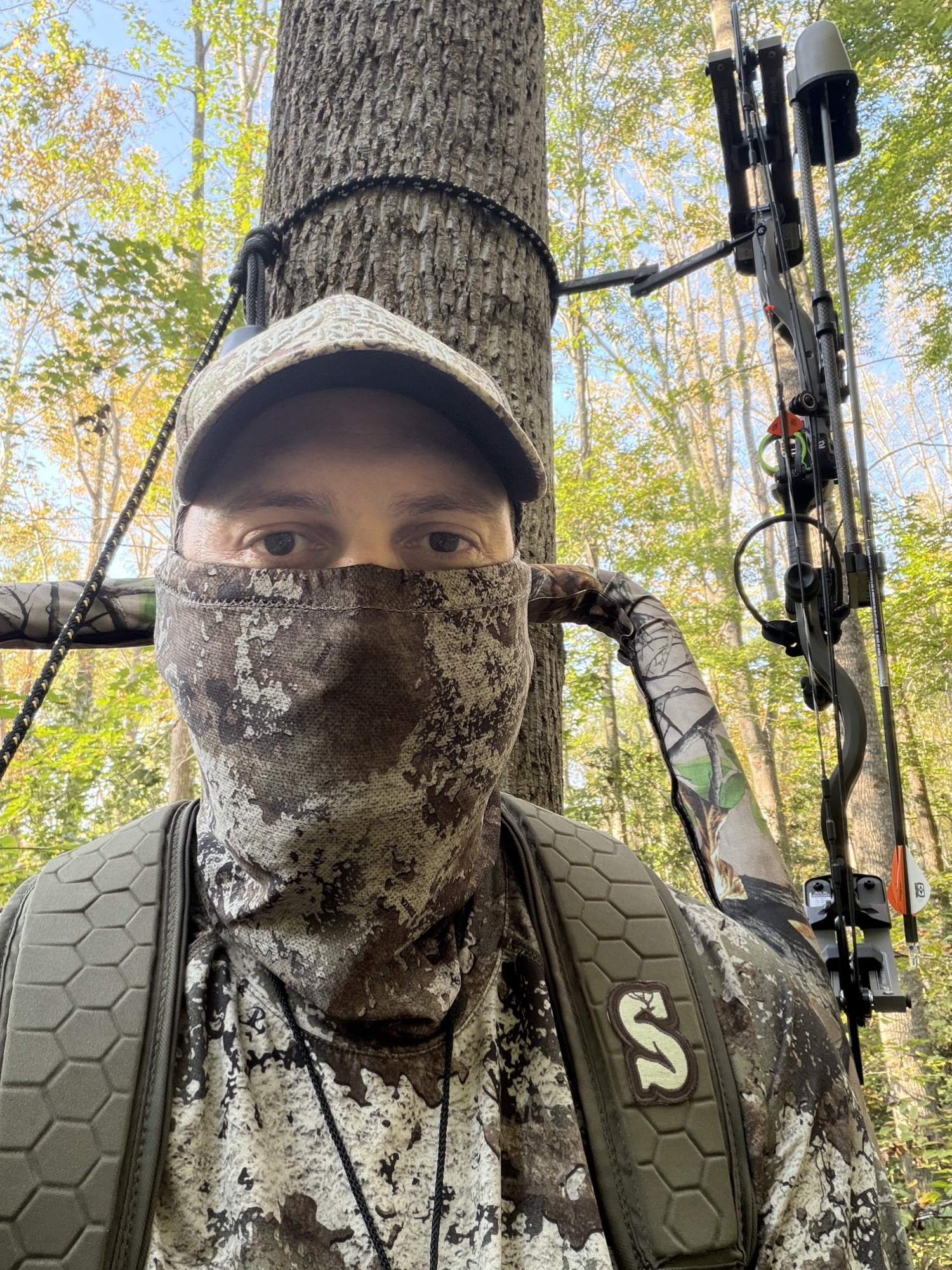 John Mobley in a tree stand wearing a camo face mask and cap, clipped to a lifeline, with his compound bow hanging beside him