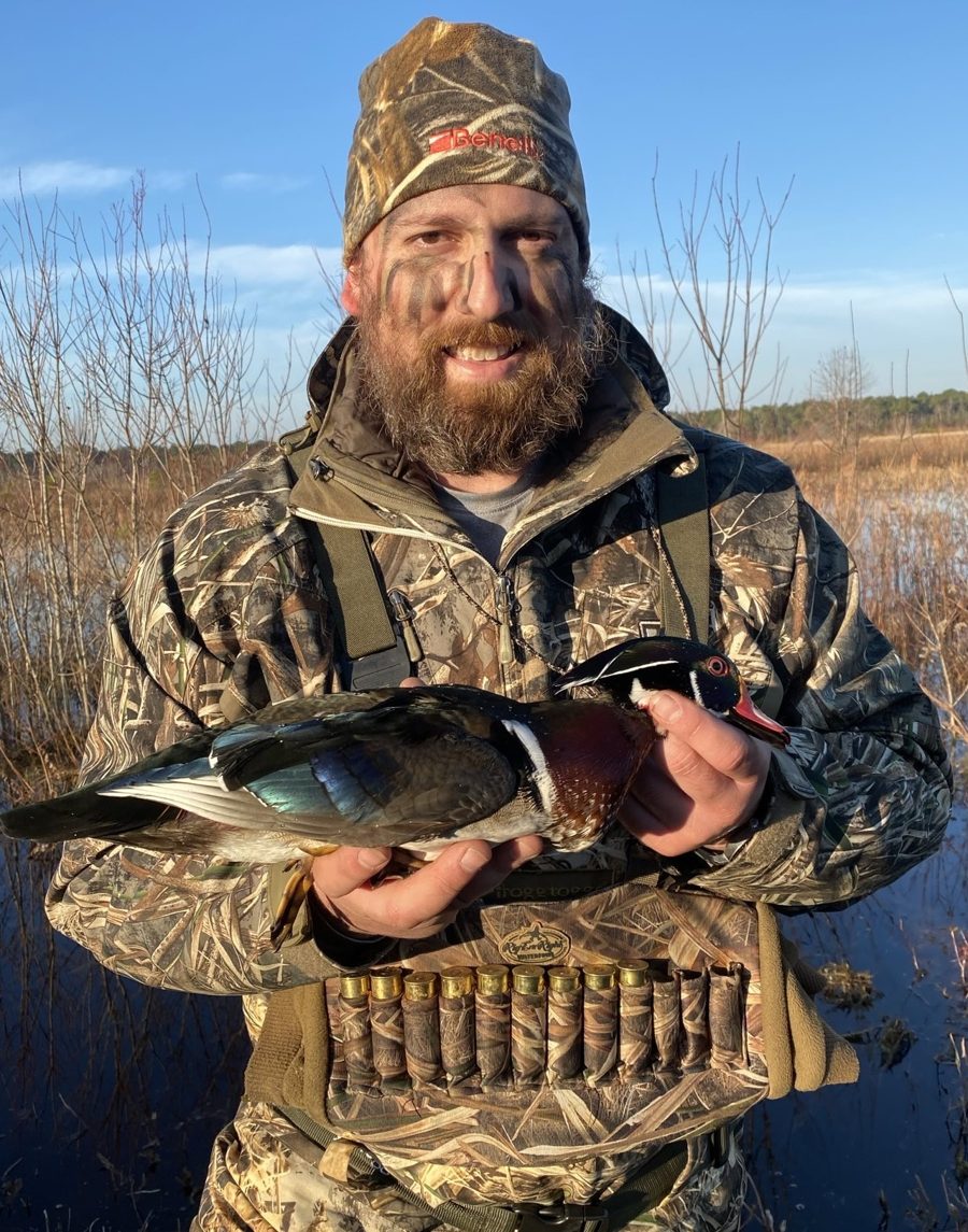 John Mobley holding a wood duck in the marsh, wearing Frogg Toggs waders and a Benelli camo beanie